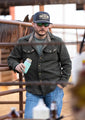 A person wearing a dark jacket, blue jeans, a Rural Cloth Longhorn Hat, and sunglasses is standing in front of a metal fence holding a beverage can. The background features a horse and some bottles, capturing the essence of the ranching lifestyle.