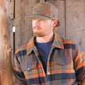 A bearded man is sporting the Rural Cloth Lumberjacket over a t-shirt and a beige baseball cap. He stands beside a wooden wall, gazing to the side.