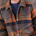 A person is wearing the Lumberjacket by Rural Cloth, a buttoned-up, Sherpa-lined jacket with a collar, featuring plaid patterns in shades of brown, orange, navy, and green. The real men’s wool polyester jacket has two chest pockets with buttons. The person is also wearing a dark blue T-shirt underneath.