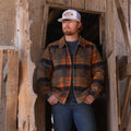 A person stands in a rustic wooden doorway wearing a Lumberjacket from Rural Cloth, jeans, and a white baseball cap labeled 