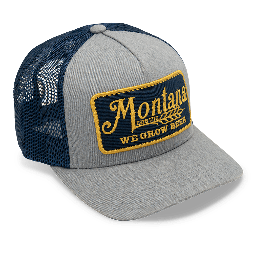 The Montana We Grow Beer Hat by Rural Cloth is a stylish gray trucker hat featuring a blue, breathable mesh back and blue brim. It sports a yellow-embroidered patch on the front that reads "Montana" with "Est. 1786" underneath, followed by "We grow beer." This hat also includes an adjustable snapback closure for a perfect fit.