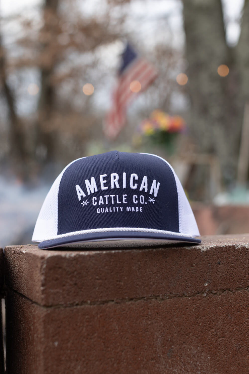 A Rural Cloth Quality Made Hat, distinguished by its navy and white trucker design and embroidered "AMERICAN CATTLE CO. QUALITY MADE" logo, rests on a brick ledge. The blurred background showcases an out-of-focus American flag amid a fuzzy outdoor setting. The hat includes a snapback closure for an adjustable fit.