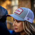 A woman with long blonde hair wears the Rc X CL Hat by Rural Cloth, a gray and white mesh baseball cap featuring a Coors Light logo and an American flag patch on the side. The hat includes a convenient snapback closure, ensuring a comfortable fit as she looks ahead, the background tastefully blurred.