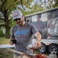 A man wearing a Rural Cloth Rc X CL Hat and sunglasses is pouring a can of Coors Light over a plate of crawfish. He stands next to a trailer with an image of someone dressed in a gray T-shirt that reads 