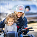 A woman wearing a gray Rc X CL Hat from Rural Cloth with a snapback closure and a dark hoodie sits on an ATV, guiding a young child in a gray shirt who is holding the handlebars. A blurred vehicle is in the background.