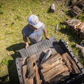 A man in a grey T-shirt and the Rural Cloth Rc X CL Hat with a snapback closure unloads firewood from the tailgate of a pickup truck. The truck bed contains a stack of cut logs, and he stands on a grassy area with some additional wood and logs scattered nearby.