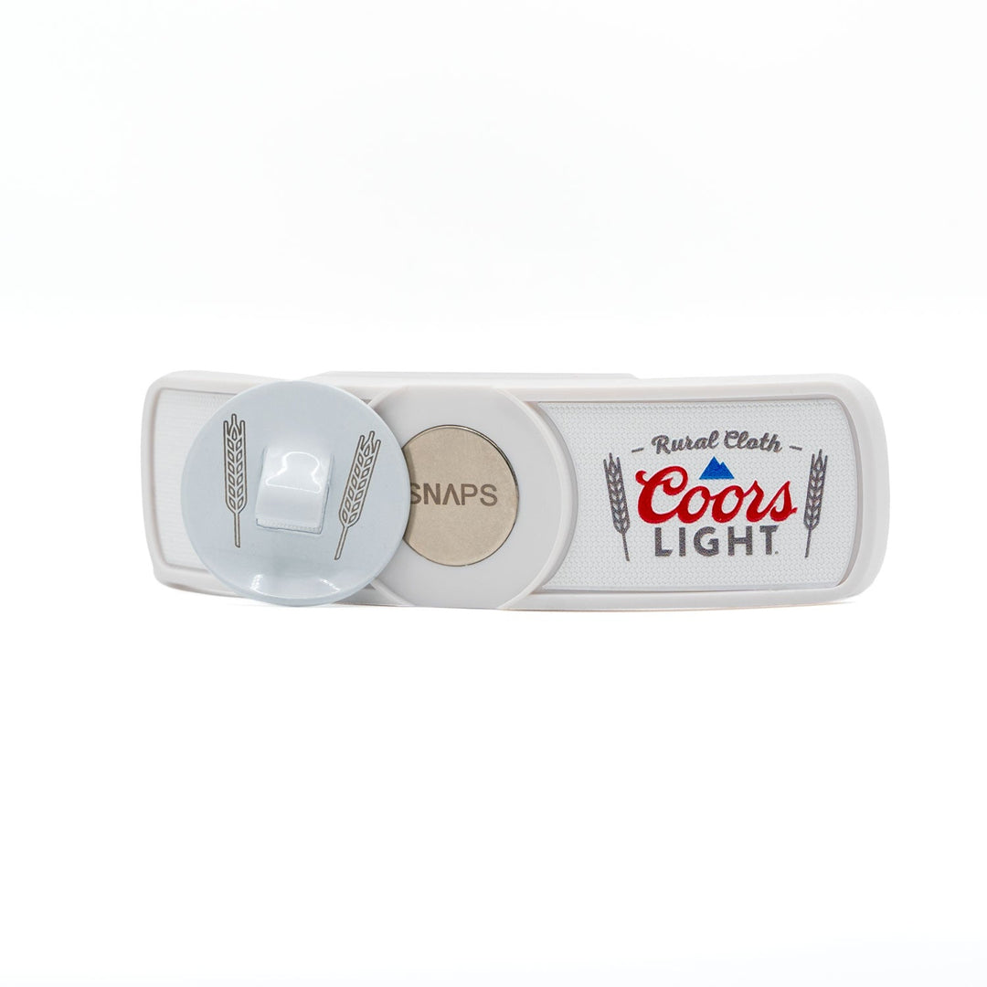 The RC x CL SNAPS Bottle Opener from Rural Cloth showcases a white belt buckle adorned with the Coors Light logo and wheat designs. It includes "SNAPS" and a convenient bottle opener positioned on the left side, making it an ideal accessory for fans of longneck drinks.