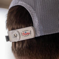 A person sporting a white mesh cap from the RC x CL SNAPS Bundle by Rural Cloth, adorned with a white plastic SNAPS Bottle Opener. The clip features labels 