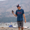 A man in a blue t-shirt and gray shorts, wearing a Retro Coors Light Hat by Rural Cloth, strolls along a rocky beach with his eyes glued to his phone. The serene backdrop showcases a lake dotted with boats and rolling hills, embodying the essence of rural living.