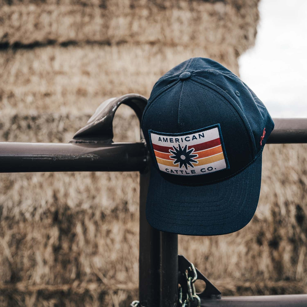 A black Retro Spur Hat from Rural Cloth, adorned with a logo reading "American Cattle Co." and featuring a cow and sun design, is hanging on a metal fence. In the background, large hay bales are stacked together.