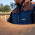 A person in the Rider quilted vest by Rural Cloth, featuring blue and brown tones, stands outdoors near a wooden fence. The focus is on their upper body and the insulated vest, with the background slightly blurred. The person appears to be interacting with an animal that is partially visible in the foreground.