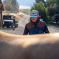 A man wearing the Rural Cloth Rider vest and a baseball cap is partially obscured by a large animal, likely a horse. He is outdoors on a dirt road with trees and an off-road vehicle in the background.