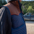 A person is shown from the shoulders down, wearing the Rider blue quilted vest with a brown faux leather yoke by Rural Cloth, along with a gray hooded sweatshirt underneath. A blurry vehicle and trees are visible in the background.