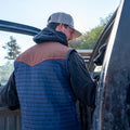 A person wearing Rural Cloth's blue and brown quilted Rider vest and a gray cap stands outside a vehicle with the door open, facing a large black dog inside the vehicle. Trees and a bright sky are visible in the background, creating a serene outdoor setting.