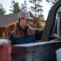 A man wearing the Rider cap and jacket by Rural Cloth, layered with a quilted insulated vest, stands beside a vehicle or machinery, intently looking down at it. There is a wooden cabin and trees in the background.