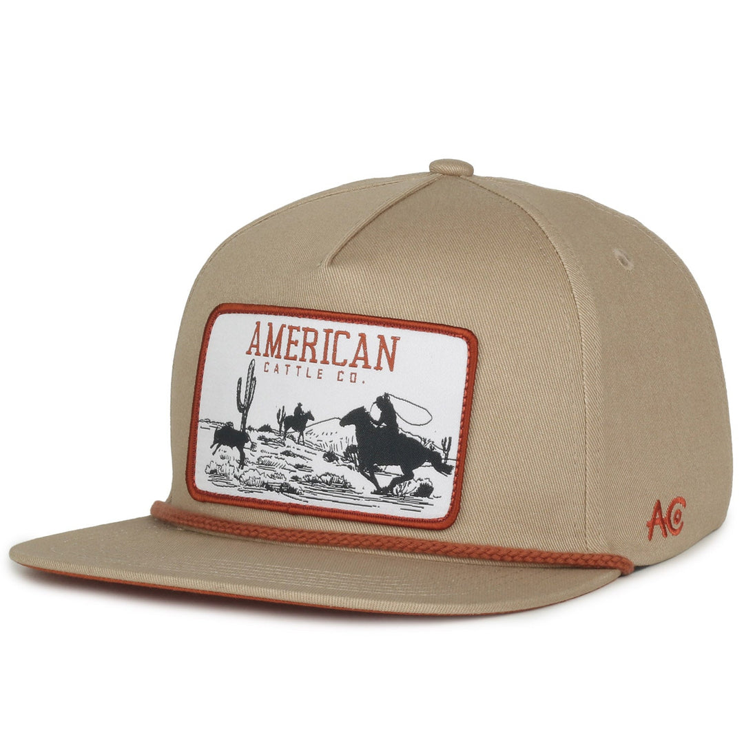 The Roper Hat by Rural Cloth is a beige baseball cap with Western flair, featuring a cowboy roping cattle among desert cacti and "American Cattle Co." at the top. A brown string along the brim completes this must-have for ranching accessory enthusiasts.