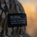 The Rural Def Bottomland Edition by Rural Cloth is a Mossy Oak Bottomland Camo Hat featuring a patch with the word 