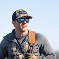 A man donning a Rural Cloth Rural Def - Bottomland Edition, reflective sunglasses, and outdoor gear stands against a clear blue sky. Embracing the rural lifestyle, he has a serious expression and carries what appears to be hunting and fishing equipment over his shoulder.