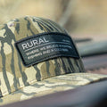 A close-up of a Rural Def - Bottomland Edition Snapback Hat by Rural Cloth with a black patch. The patch reads: 