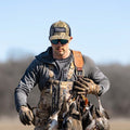 A person wearing camouflage clothing, a Rural Def - Bottomland Edition Snapback Hat from Rural Cloth, and sunglasses is carrying several birds by their feet in an outdoor setting. The background features a clear sky and blurred trees, capturing the essence of the rural lifestyle.