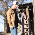 A bearded man wearing a baseball cap, black shirt, and the Sunday Jacket by Rural Cloth‚Äîa beige quilted shirt jacket with adjustable cuffs‚Äîstands outdoors holding onto a metal gate, with a wooden structure and trees in the background.