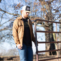 A bearded person stands next to a metal fence outdoors, surrounded by leafless trees. They are wearing the Sunday Jacket by Rural Cloth, which features a brown quilted design, Polar Fleece lining, and adjustable cuffs for added comfort. Completing the outfit are blue jeans and a white baseball cap, while they look down with a neutral expression.