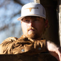 A bearded man with a serious expression wears a white baseball cap emblazoned with a golden bull emblem and the Sunday Jacket from Rural Cloth, featuring Polar Fleece lining. He leans on a wooden surface outdoors, with blurred trees visible in the background.