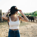 A woman wearing a white tank top and blue jeans adjusts her black Sunrise Hat by Rural Cloth. She has long hair and visible tattoos on her left arm. In the background, there are cows and a wooden fence in a rural setting.