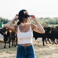 A person wearing a white tank top, blue jeans, and an adjustable Sunrise Hat by Rural Cloth stands in front of a herd of cows. The individual is adjusting their cap with both hands, revealing tattoos on their arms. The background showcases a picturesque rural landscape with trees and fields.