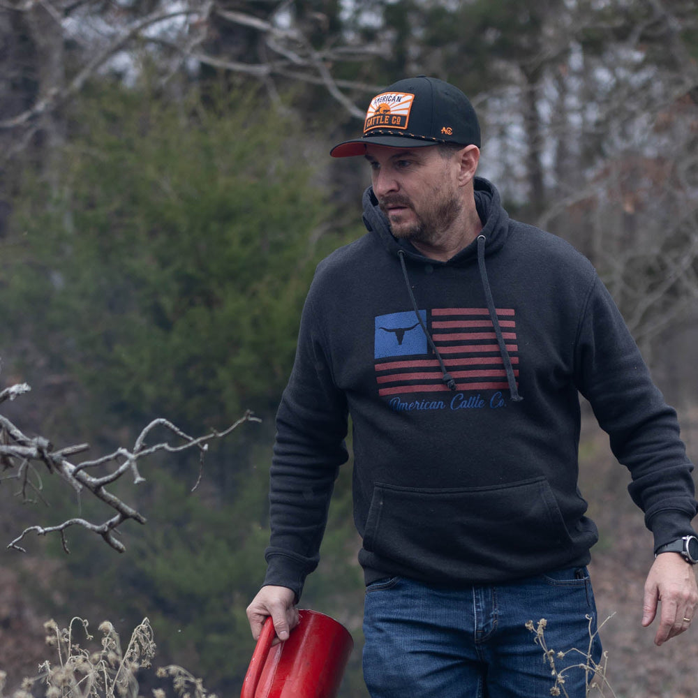 A man in a dark hoodie with an American flag design and wearing the Rural Cloth Sunrise Hat holds a red object. He is outdoors in a wooded area, with dry plants and bare trees in the background. His expression is focused.