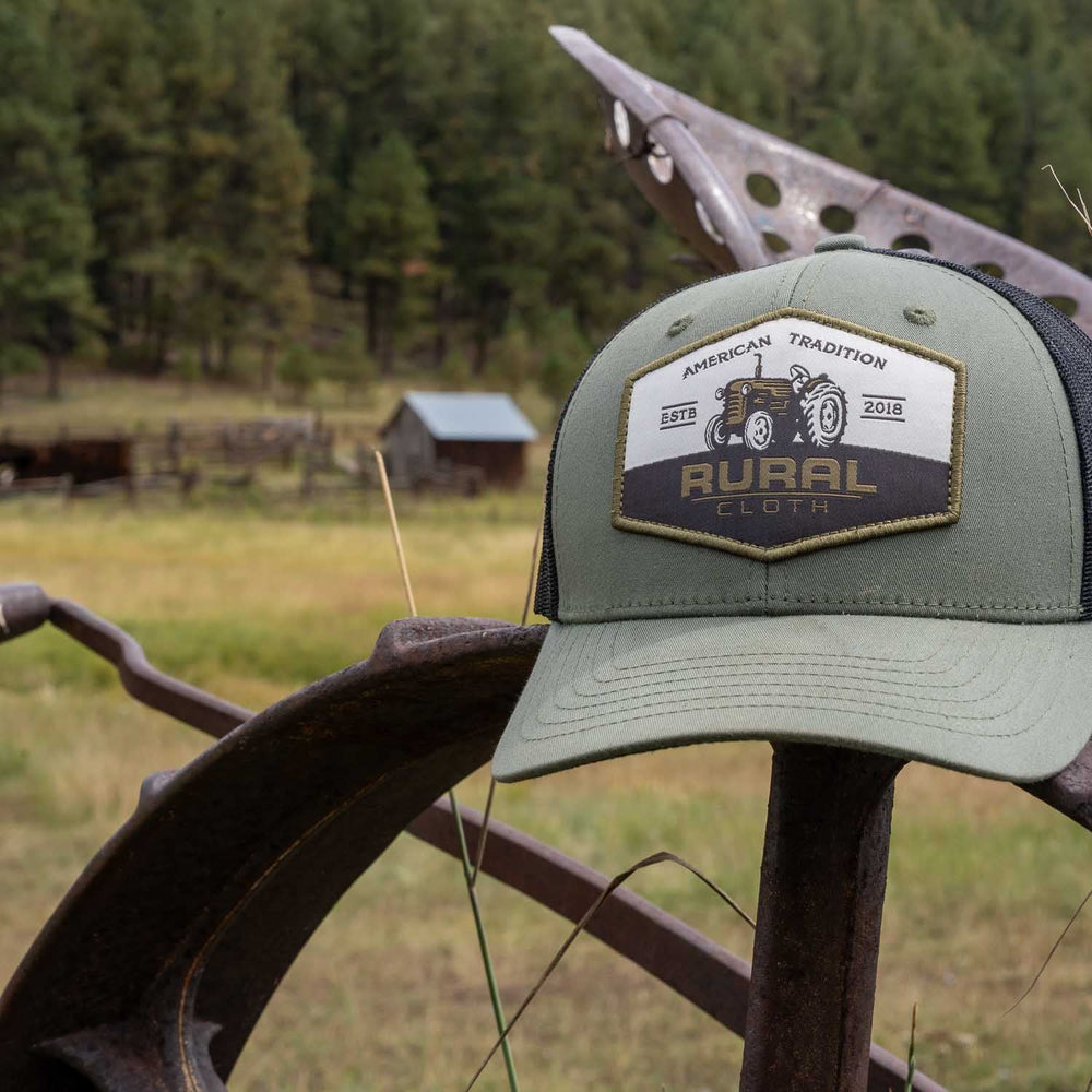 A green and black Tractor Hat from Rural Cloth, featuring breathable mesh and a patch that reads "RURAL CLOTH American Tradition," rests on the rusted iron wheel of farm machinery. In the background, an open field, an old wooden structure, and a forested area are visible.