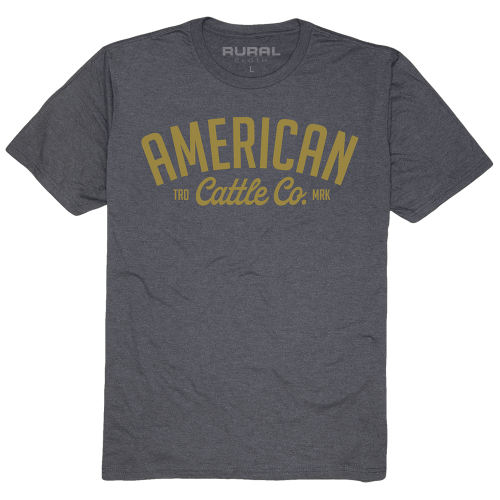 A dark gray Trademark Tee by Rural Cloth, crafted from premium weight fabric, features the text "American Cattle Co." in large yellow letters on the front. Above "Cattle Co.", smaller text reads "TRD MRK." The inner neck label displays the brand name "Rural Cloth.