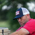 A man wearing a Rural Cloth United We Stand Hat and a red shirt is focused on an activity outdoors. He has light facial hair and appears to be in a natural, rural American setting with a green, blurred background.