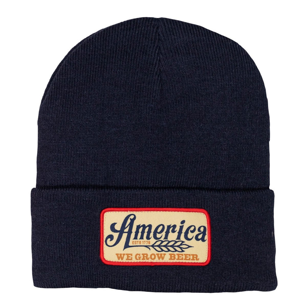 The "We Grow Beer Beanie" by Rural Cloth is a navy knit hat featuring a rectangular patch. The patch displays the word "America" in blue and "WE GROW BEER" in red, with an accompanying small wheat design. A red border around the patch accentuates America's beloved beverage on this fashionable beanie.