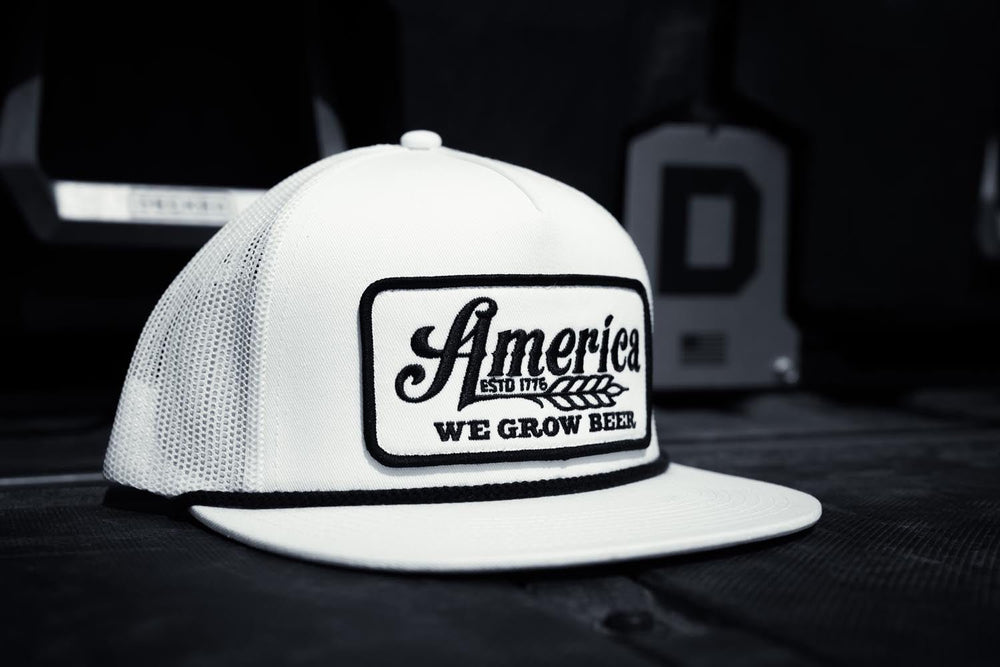 The We Grow Beer Flat by Rural Cloth is a stylish white baseball cap featuring "America's" and "We Grow Beer" text with a wheat graphic on the front. This high-profile, 5-panel flat bill hat includes a breathable mesh back and convenient snapback closure. It rests on a dark surface with a blurred background enhancing its presentation.