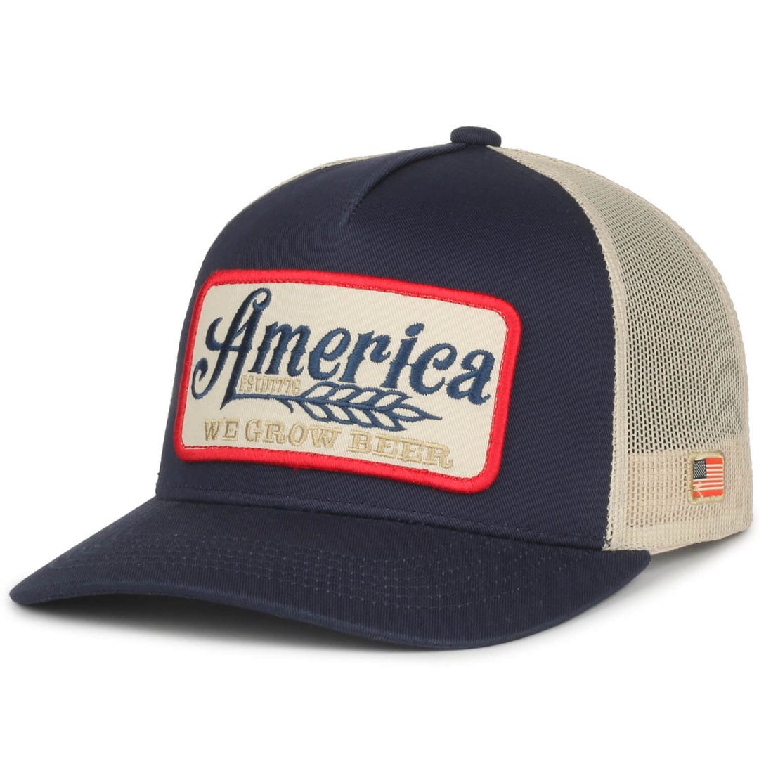 The We Grow Beer Hat-Banquet Heavy from Rural Cloth is a stylish navy blue and beige mesh trucker hat featuring a front patch that reads "America We Grow Beer" alongside a wheat graphic. The patch is bordered in red and complemented by a small American flag patch on the side. This American-made hat also features an adjustable snapback closure for a perfect fit.