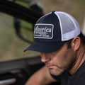 A man is seen sitting in a vehicle, wearing a blue and white mesh baseball cap with a logo on the front that reads, 