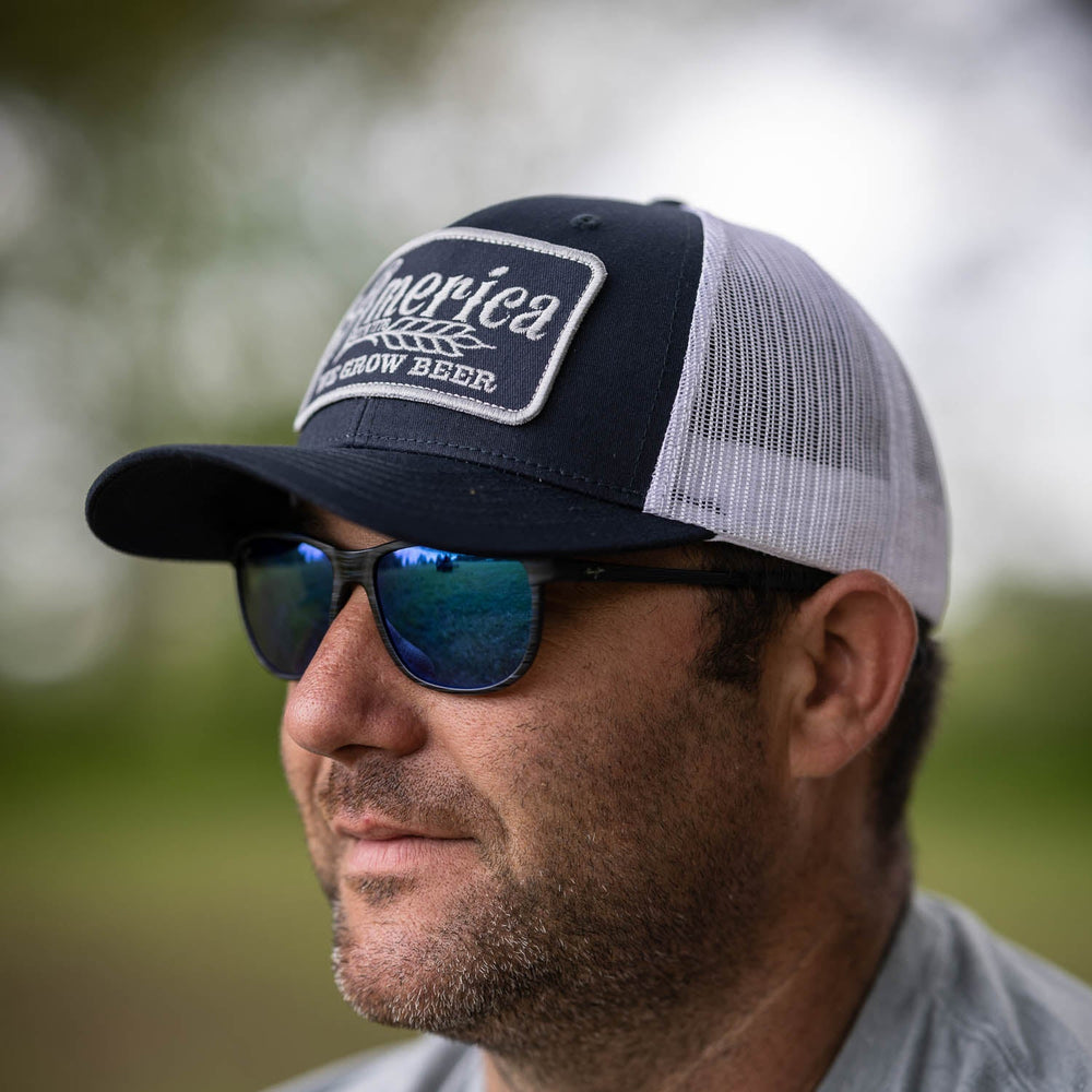 A man with stubble is wearing a navy and white We Grow Beer Hat-Blue/White by Rural Cloth, featuring an embroidered patch and reflective sunglasses. The breathable mesh back offers comfort, and the adjustable snapback closure ensures a perfect fit. The background is slightly blurred, indicating an outdoor setting.