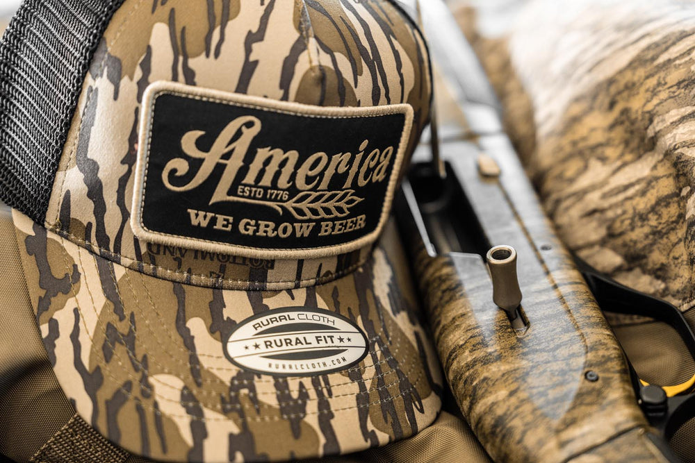 A "We Grow Beer Hat-Bottomland" camouflage snapback hat by Rural Cloth, featuring the text "America ESTD 1776 WE GROW BEER" and a "RURAL FIT" sticker, lies next to a Mossy Oak Bottomland camo shotgun on a matching patterned surface, perfectly capturing the essence of rural lifestyle.