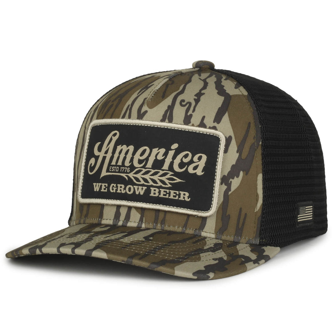 The We Grow Beer Hat-Bottomland by Rural Cloth is a camouflage trucker hat with a black mesh back and a front patch that reads "America ESTD 1776" in cursive above an illustration of wheat, with "WE GROW BEER" written below. It also features a small American flag on the side, making it perfect for embracing the rural lifestyle with its Mossy Oak Bottomland pattern.