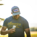 A man is holding a can of beer while looking down. He is wearing a We Grow Beer Hat-Gray by Rural Cloth, which features an adjustable snapback closure and a breathable mesh back. Additionally, he sports a blue T-shirt with yellow lettering that says 