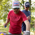 Two men are outdoors; one is grilling in a red 