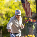 A man wearing a We Grow Beer Hat-Gray from Rural Cloth and sunglasses tends to a barbecue, holding a can of Coors Light beer in one hand. An American flag is visible in the background, and bright greenery surrounds the scene.