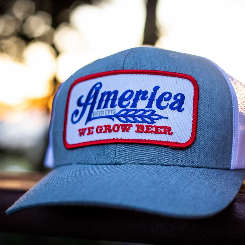 The We Grow Beer Hat-Gray by Rural Cloth features a rectangular patch showcasing the word "America" in blue cursive font over an image of wheat, with the slogan "We Grow Beer" in red. This gray and white trucker hat includes an adjustable snapback closure and a breathable mesh back for comfort, all set against a blurred outdoor background.