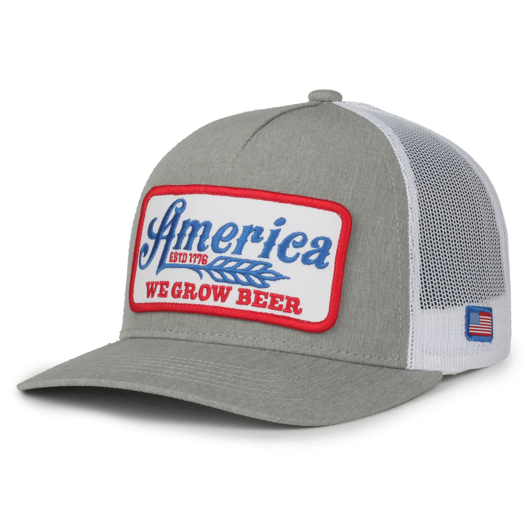The We Grow Beer Hat-Gray by Rural Cloth is a gray and white trucker hat with a mesh back. Its front showcases a patch with "America" in blue, "ESTD 1776" in red, a wheat design, and "WE GROW BEER" in red letters. An American flag patch adorns the side. This hat features an adjustable snapback closure and is proudly made in the USA.