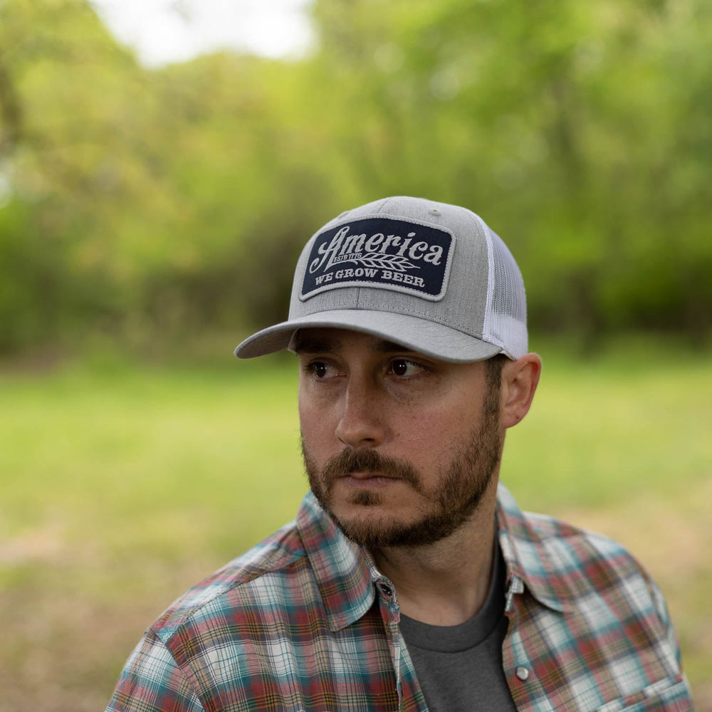 A man with a beard is wearing a plaid shirt and the Rural Cloth "We Grow Beer Hat-Gray/Blue," which features an adjustable snapback closure and a breathable mesh back. He is outdoors with a blurred background of greenery, looking to the side.