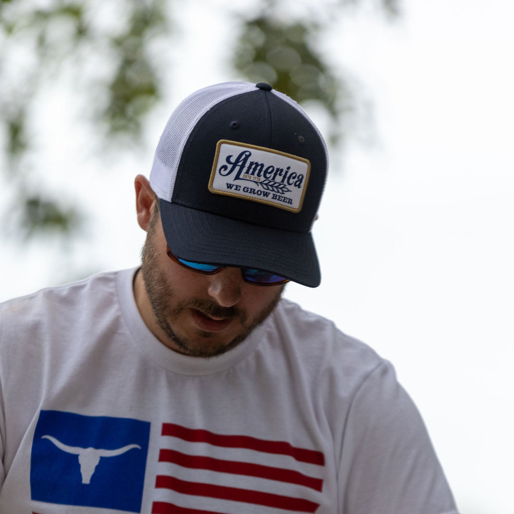 A man wearing a white and blue "We Grow Beer Hat-Less Filling!" cap from Rural Cloth with an adjustable snapback closure and a white shirt featuring a Texas Longhorn logo over red and blue stripes. He is looking downwards, with sunglasses on, and trees blurred in the background.