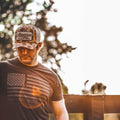 A man stands outdoors, leaning on a wooden railing while wearing a We Grow Beer Hat in Old School Camo by Rural Cloth. He sports a dark t-shirt featuring an American flag and the words 