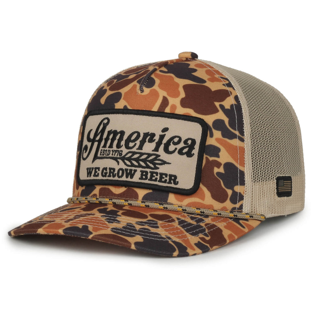 The We Grow Beer Hat-Old School Camo by Rural Cloth is a camouflage trucker hat featuring a front panel with the embroidered patch text "America ESTD 1776 WE GROW BEER." It includes a mesh back and an adjustable snap closure, making it ideal for fans of retro country music.