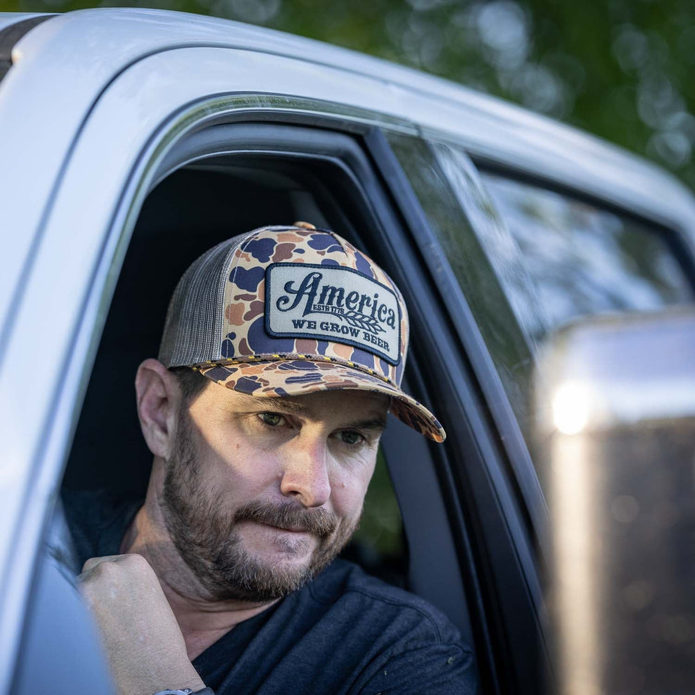 A man in a vehicle is gazing out of the open driver's side window. Wearing his cherished Rural Cloth "We Grow Beer Hat-Old School Camo," his right hand rests near his chin in thoughtful contemplation. Trees are visible in the background, adding a serene touch to the retro country music vibe.